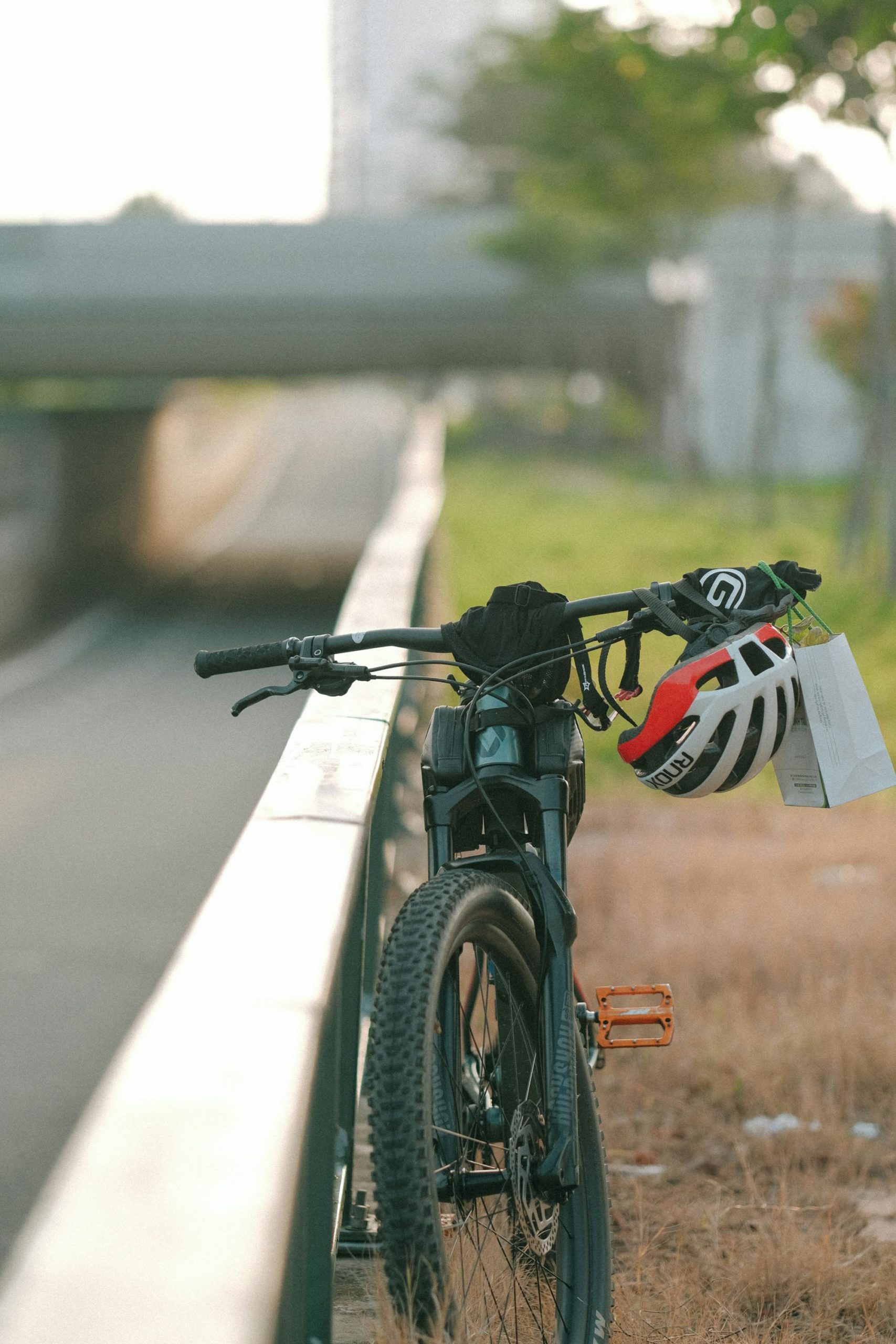A bicycle with a helmet hanging on the handlebar, parked on a road barrier in an urban setting.